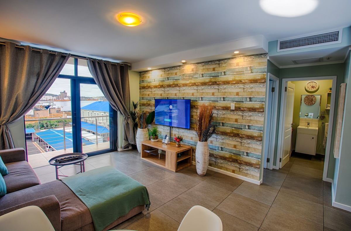 A modern living space is highlighted by large windows that offer harbor views and natural light. The room features a cozy seating area with a sofa, a flat-screen TV mounted on a wooden accent wall, and a dining table in a corner. A doorway leads to a bathroom.