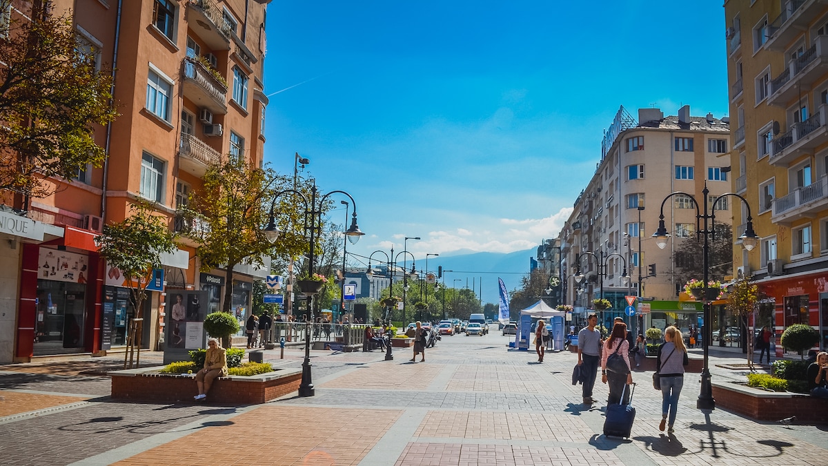 A vibrant city street is captured, showcasing pedestrians walking along the bustling promenade. Shops and cafes line both sides, while lampposts and trees add to the urban atmosphere. The backdrop features mountains, indicating a scenic horizon beyond the cityscape under a clear blue sky.