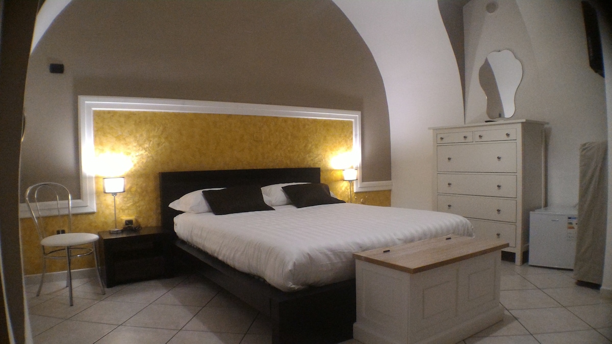 A spacious bedroom is featured with a centered bed and decorative yellow wall behind it. Flanking bedside lamps provide soft illumination. A minimalist dresser stands against one wall, while a mini fridge is discreetly placed near the bed.