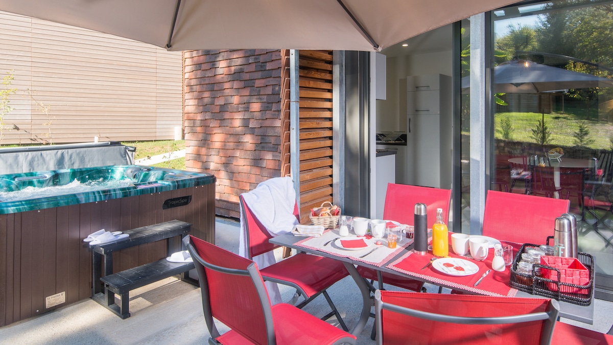 An outdoor terrace is equipped with a hot tub and a dining area. A table is set with a red tablecloth, plates, and beverages. Surrounding chairs provide ample seating, while the background features wooden paneling and greenery.