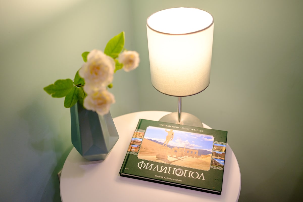 A bedside table is adorned with a modern lamp casting soft light, accompanied by a geometric vase containing delicate white flowers. A book titled 'Филипопол' rests on the table, offering insights into the local history and culture.
