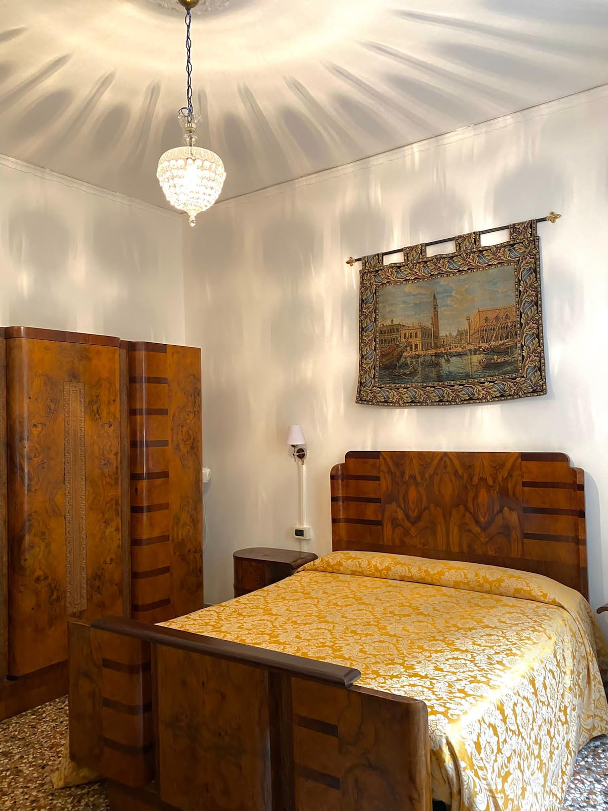 The room features vintage Venetian-style furnishings, including a spacious bed with a gold-toned coverlet. An ornate chandelier casts patterned light across the walls, complemented by a tapestry depicting a city scene. A wooden wardrobe is also visible, adding to the room's classic charm.
