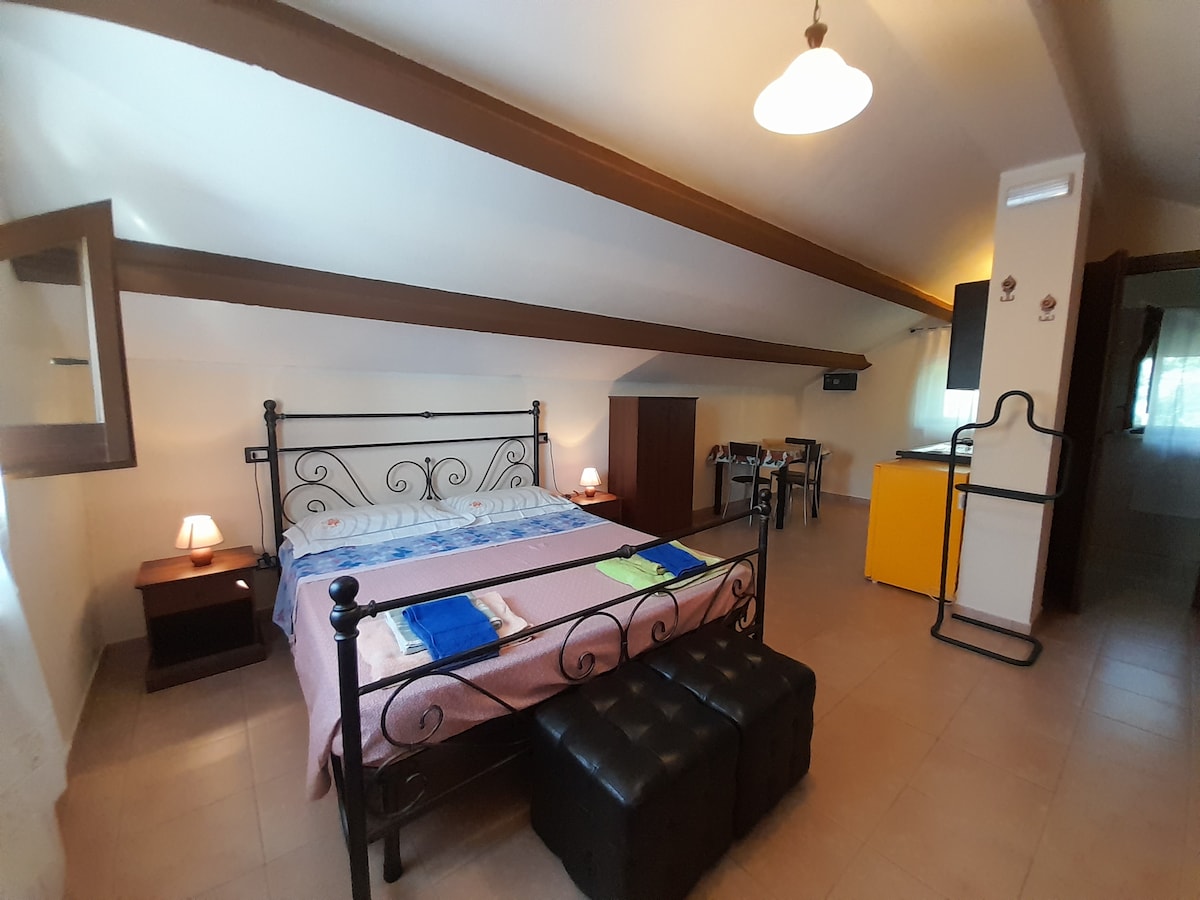 A spacious room features a wrought-iron bed dressed with colorful linens. Two bedside lamps provide soft lighting. An open kitchenette is seen in the background beside a small dining table. Dark wooden beams stretch across the ceiling, enhancing the room's openness.