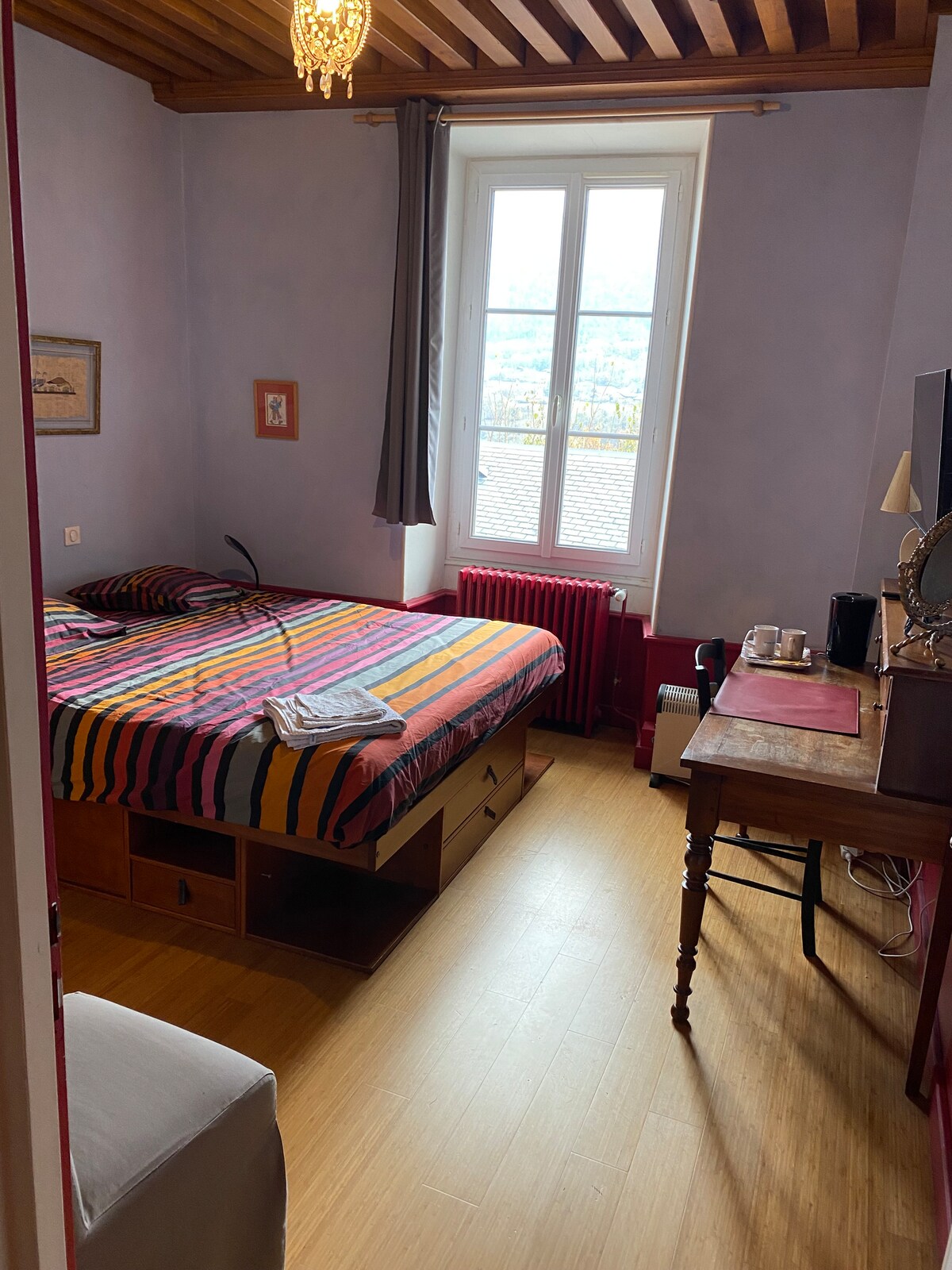 A cozy room features a brightly striped bedspread on a spacious bed, complemented by warm wooden accents. Natural light enters through a large window, illuminating a wooden desk and chair. A vintage mirror and other decorative elements add character to the inviting space.