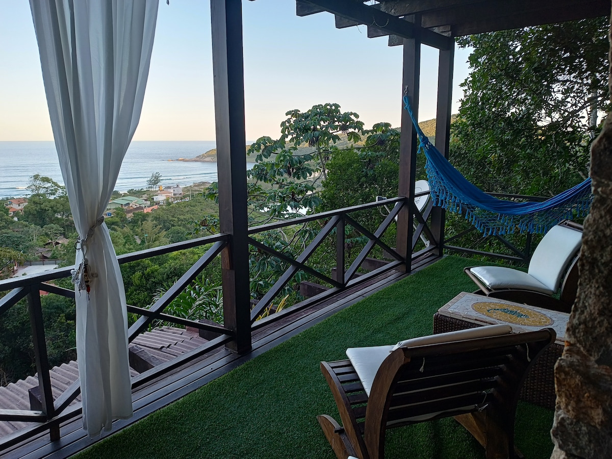 A private terrace features a view of the ocean, framed by greenery. Comfortable seating options include a hammock and a wooden chair. Light curtains billow gently in the breeze, enhancing the serene ambiance of the space while creating a connection with the outdoor surroundings.