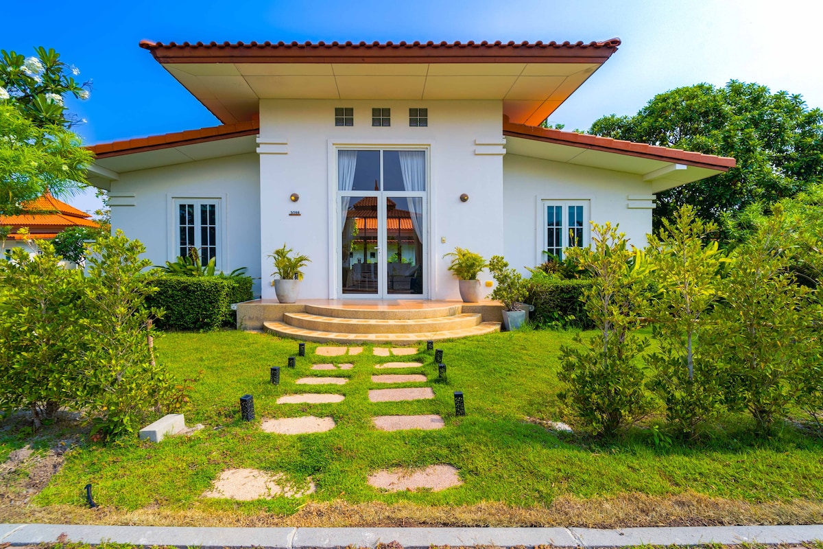 The villa's exterior features a modern design with a spacious entrance and stone steps leading to double glass doors. Lush green gardens and neatly trimmed hedges border the pathway, while a vibrant blue sky complements the inviting structure.