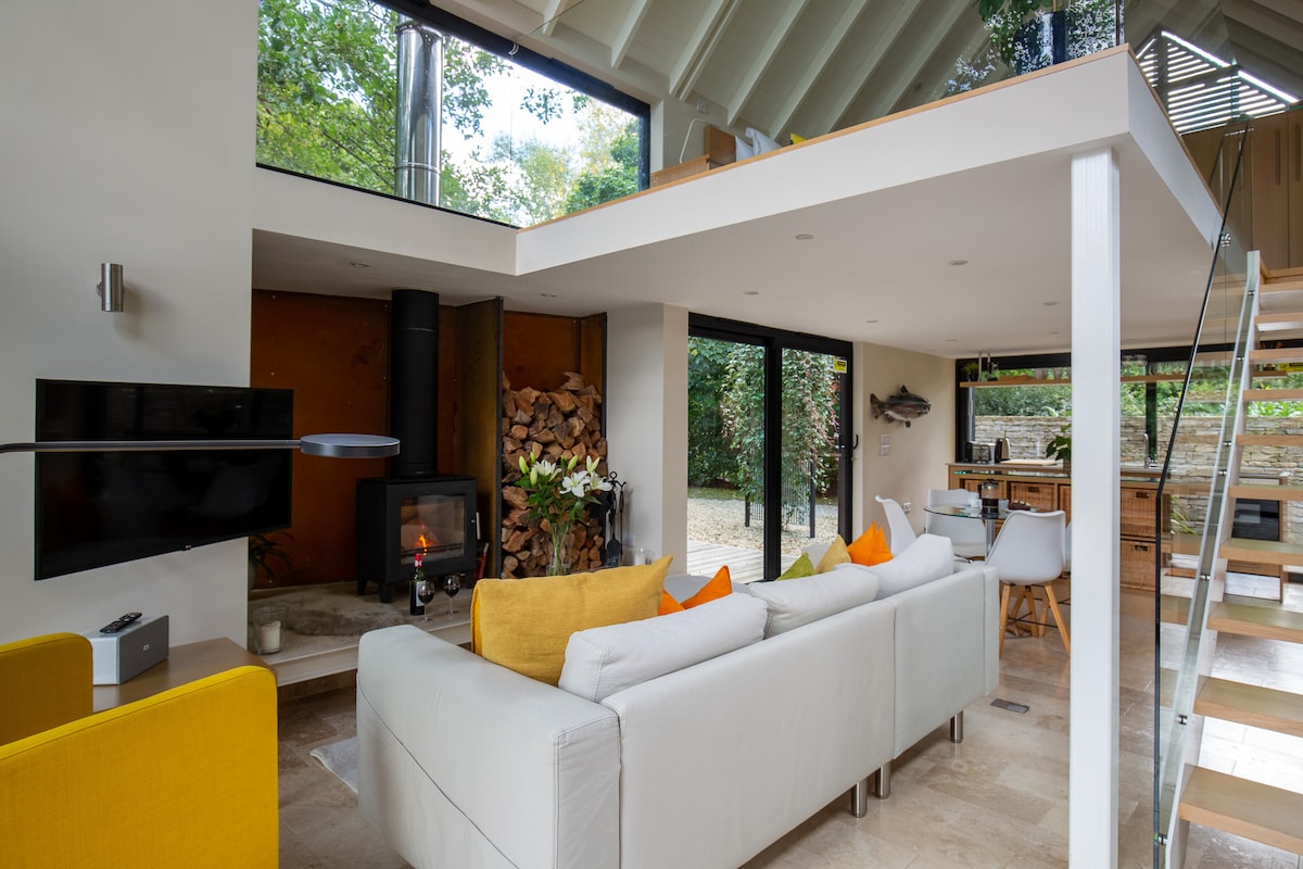 A modern living area is presented with a bright white sofa adorned with colorful cushions. A log burner is visible, surrounded by neatly stacked firewood. Large glass doors invite natural light and offer views of the outdoor space, creating a seamless connection with nature.