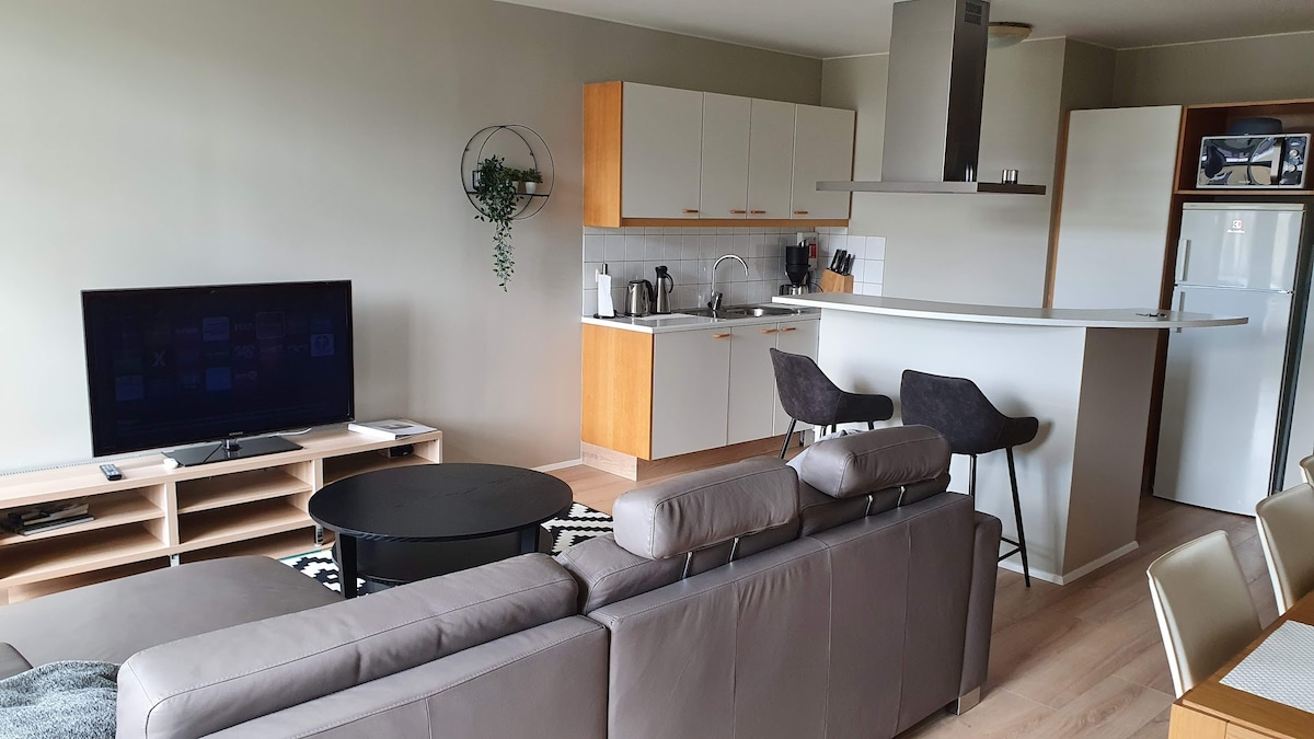 A spacious living area features a comfortable sofa and a flat-screen TV, positioned next to a stylish kitchen. The well-equipped kitchen showcases modern appliances, including a refrigerator, and is complemented by wood cabinetry. A round table sits nearby, providing a functional dining space.