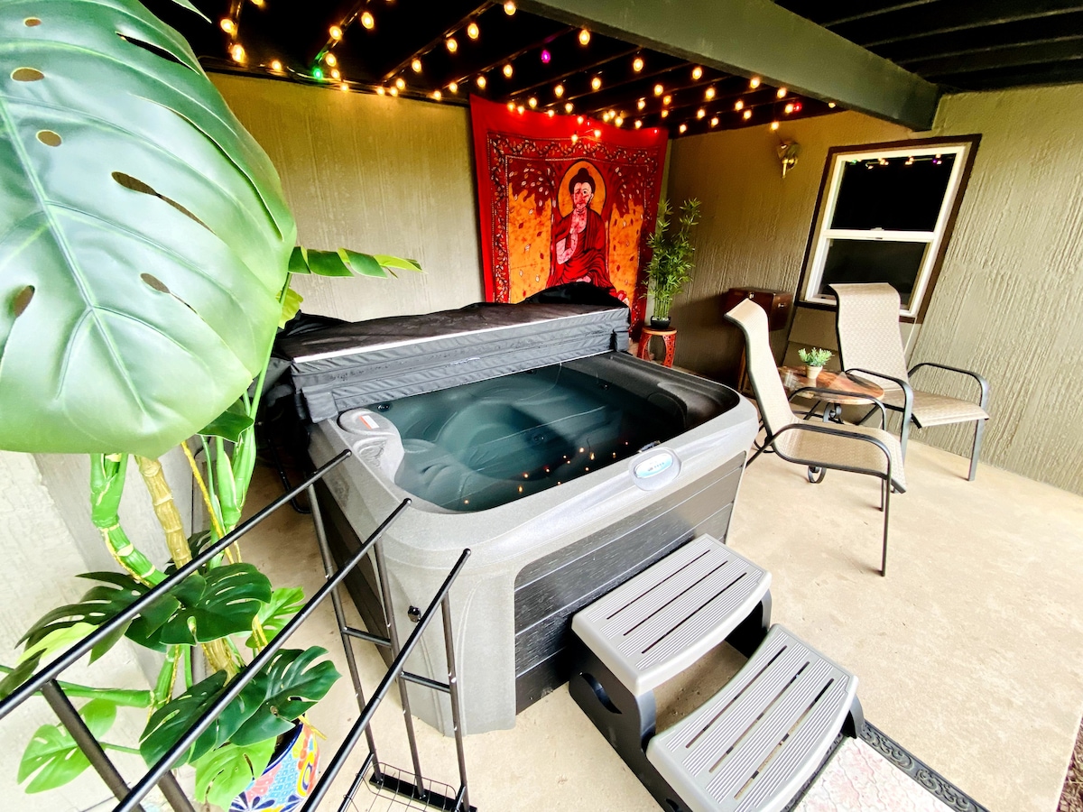 A private outdoor area features a hot tub surrounded by a leafy plant and softly lit with string lights. A small table and two chairs offer a cozy space for relaxation, with a colorful wall hanging adding a warm touch to the ambiance.