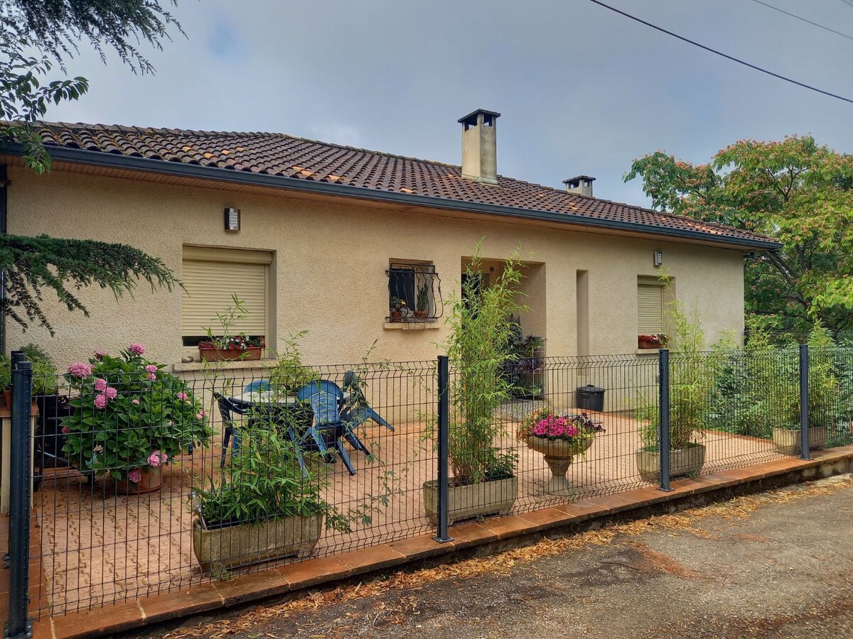 A single-story house is surrounded by a low fence, featuring a covered terrace with potted flowers and greenery. A set of chairs and a table is positioned on the patio, creating a space for relaxation. The roof is tiled, and window shutters complement the exterior.