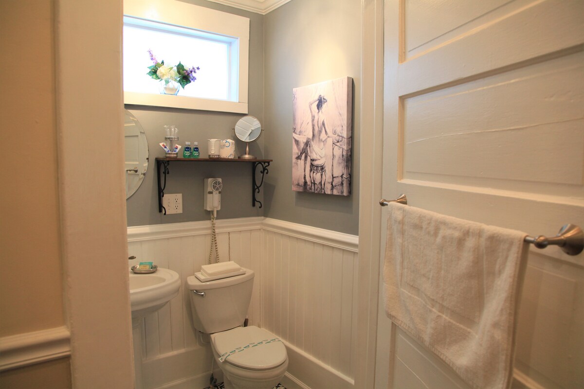 A well-appointed bathroom features light gray walls and wainscoting. A compact toilet sits beside a small shelf holding toiletries. A round mirror and a picture hang above, while a white towel is neatly hung on a bar next to the door.