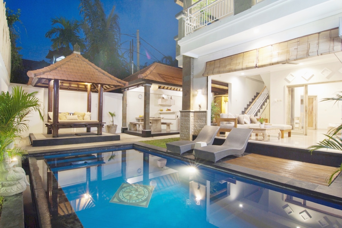 A private swimming pool reflects the evening sky, surrounded by a lush garden. A gazebo with comfortable seating is visible to the left, while a spacious outdoor area features sun loungers. The contemporary villa design includes glass doors that open to a well-lit living space.