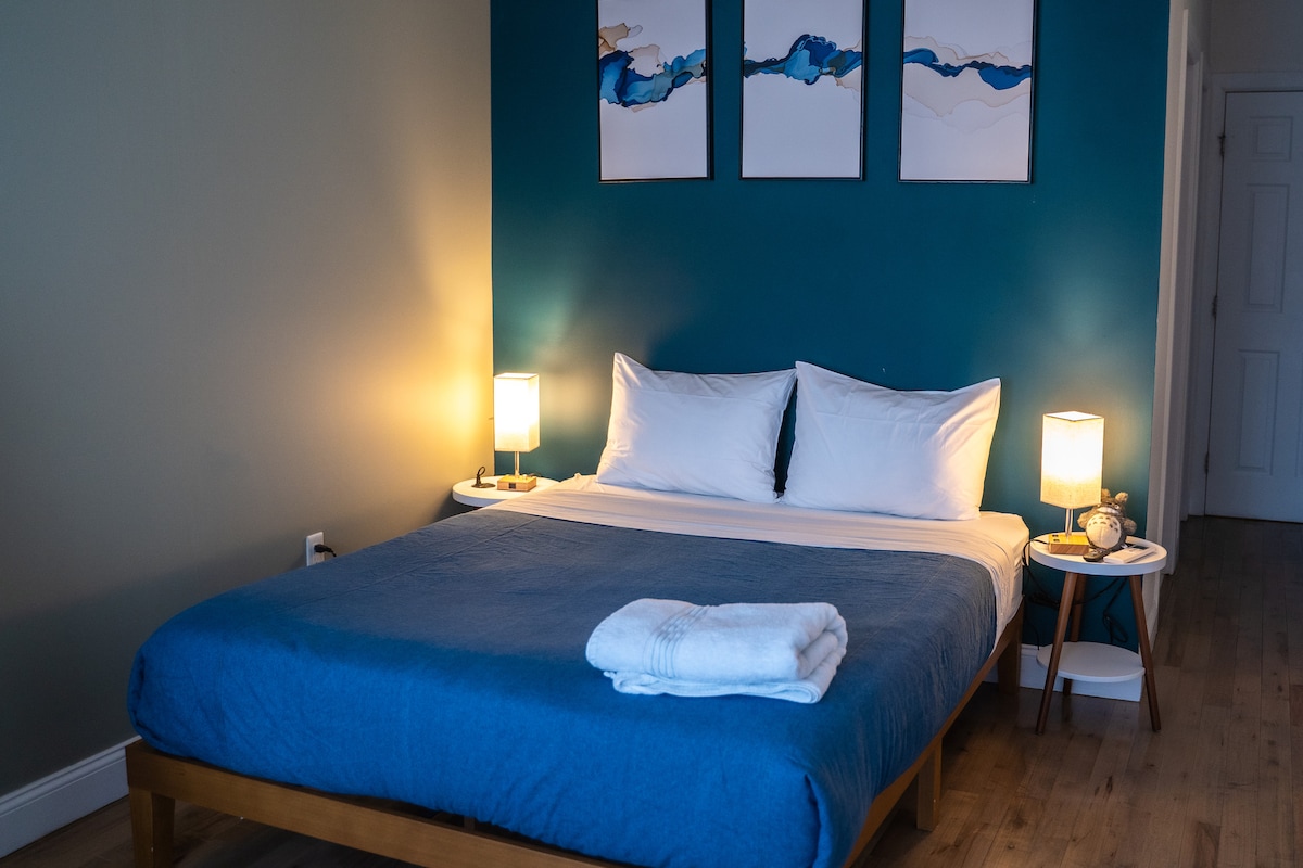 A cozy sleeping area features a spacious bed dressed in a blue blanket, complemented by crisp white pillows. On either side, nightstands with lamps provide soft lighting. Above the bed, three abstract artworks in blue and gold are displayed against a teal accent wall.