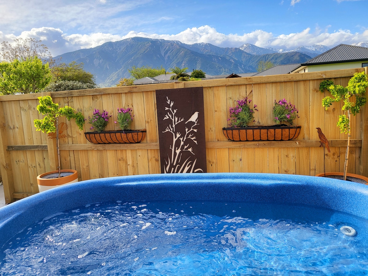 A hot tub is positioned in a wooden enclosure, surrounded by planters with purple flowers and a decorative panel featuring a bird design. The backdrop presents a scenic view of mountains under a clear sky, contributing to a serene outdoor ambiance.