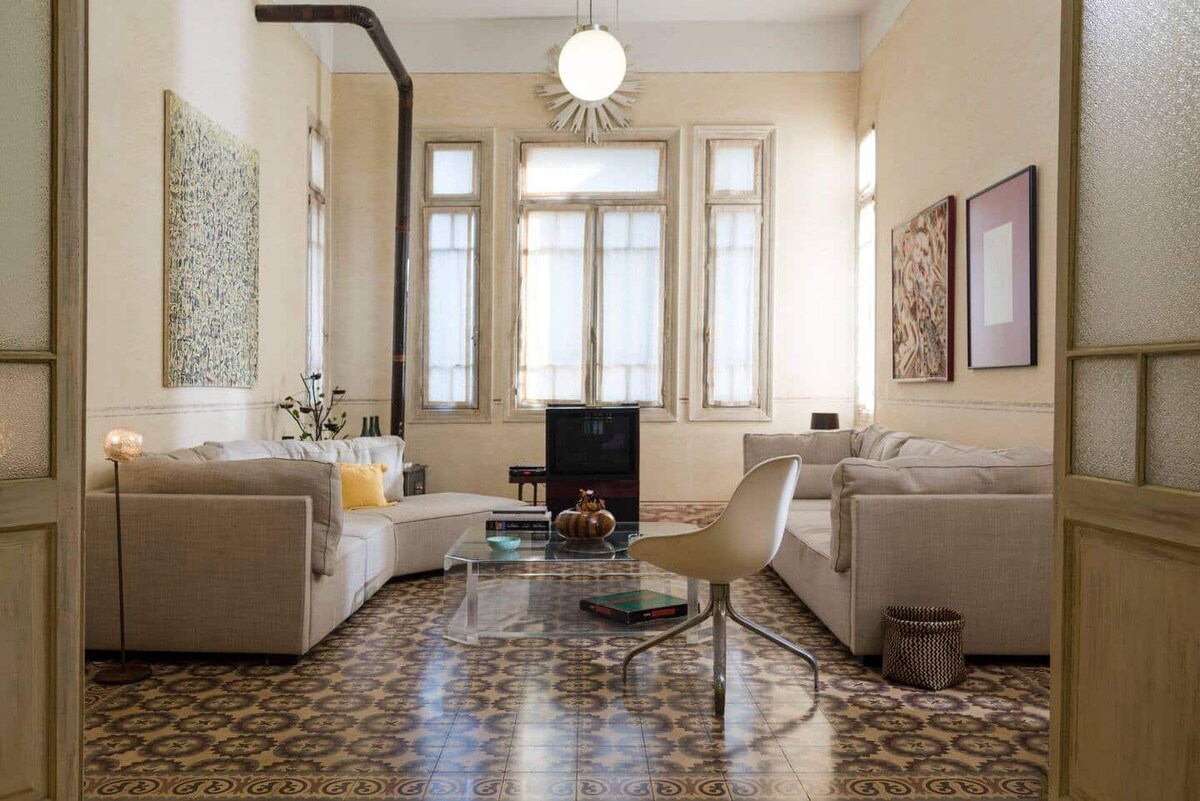 A spacious living room features large windows that allow natural light to fill the space. The area is furnished with two beige sofas and a glass coffee table. Decorative wall art adds a touch of character, while patterned floor tiles enhance the room's aesthetic.