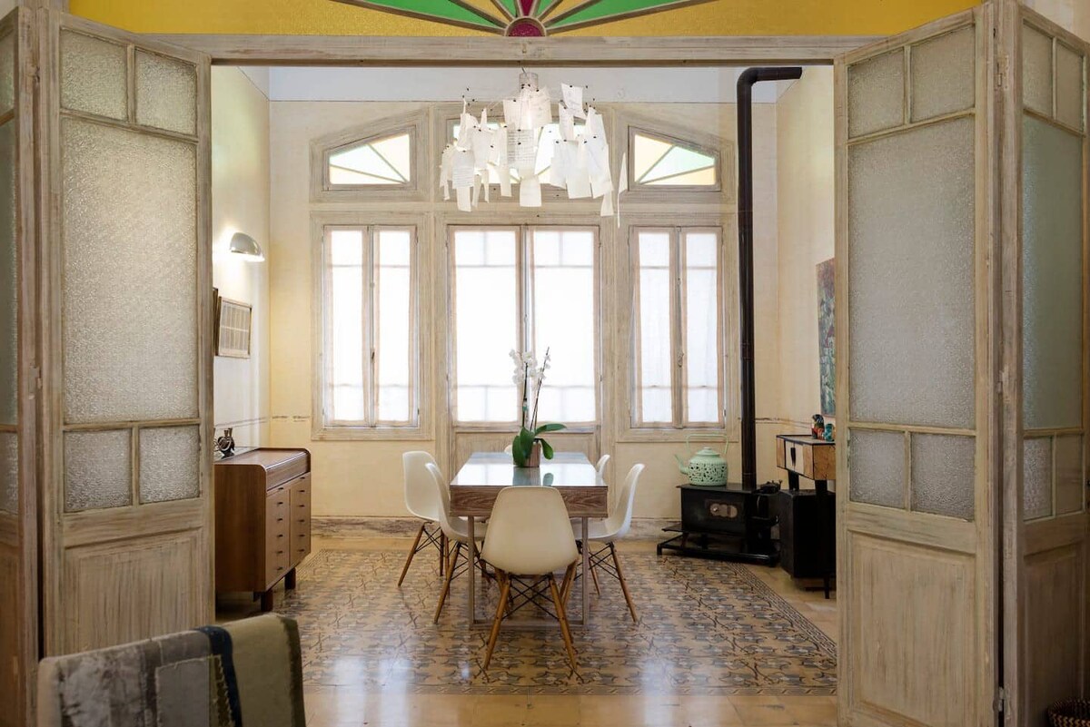 A dining area is revealed through elegant double doors, featuring a wooden table surrounded by white chairs. Large windows allow natural light to fill the space, enhancing the intricate tiled floor. The room is complemented by a stylish light fixture and decorative elements.