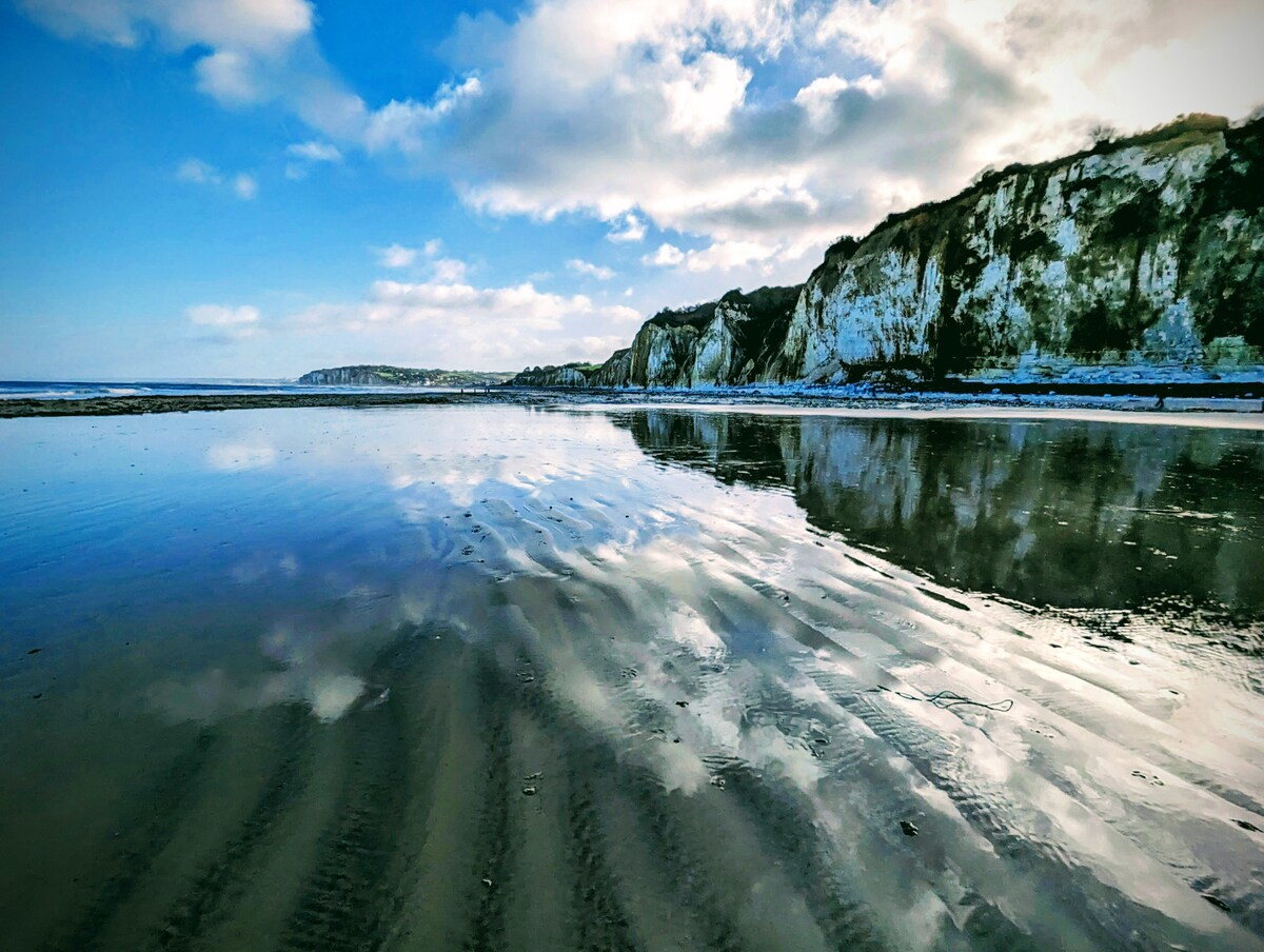 A sandy beach is shown at low tide, reflecting the sky filled with soft clouds. The rugged coastline with cliffs is visible in the distance, providing a natural backdrop to the tranquil shoreline.