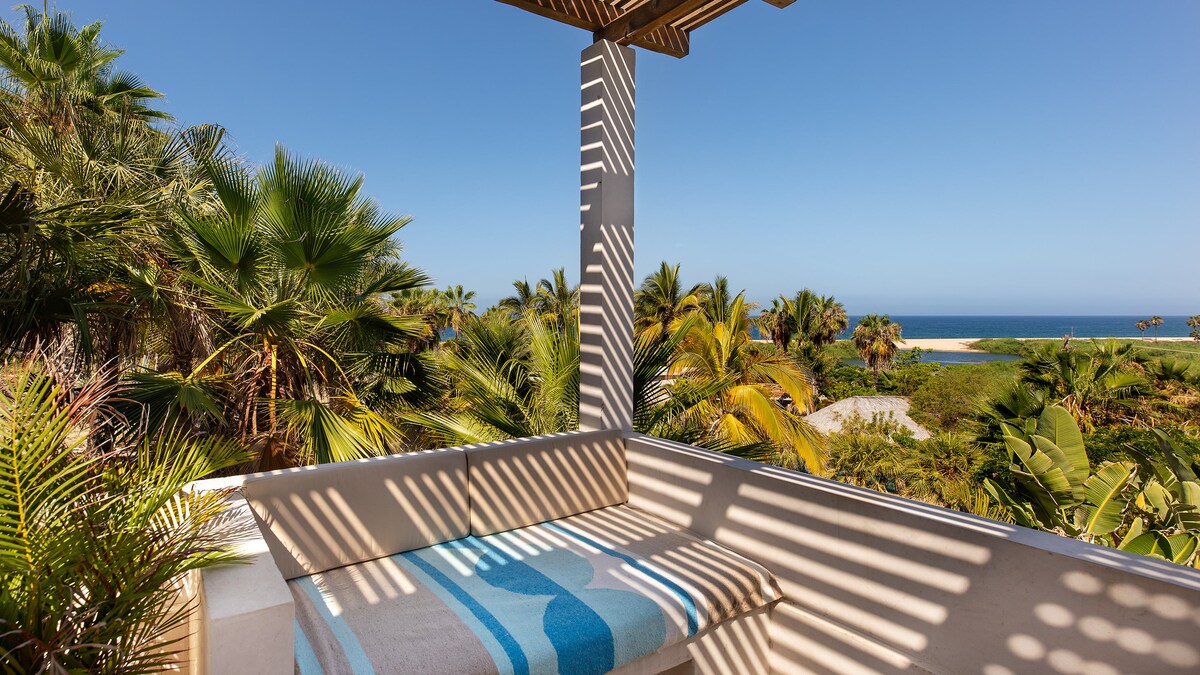 A shaded lounge area features a comfortable bench adorned with a striped blanket. Sunlight creates patterns through the slatted roof, illuminating surrounding palm trees and offering views of a serene coastline and ocean beyond.