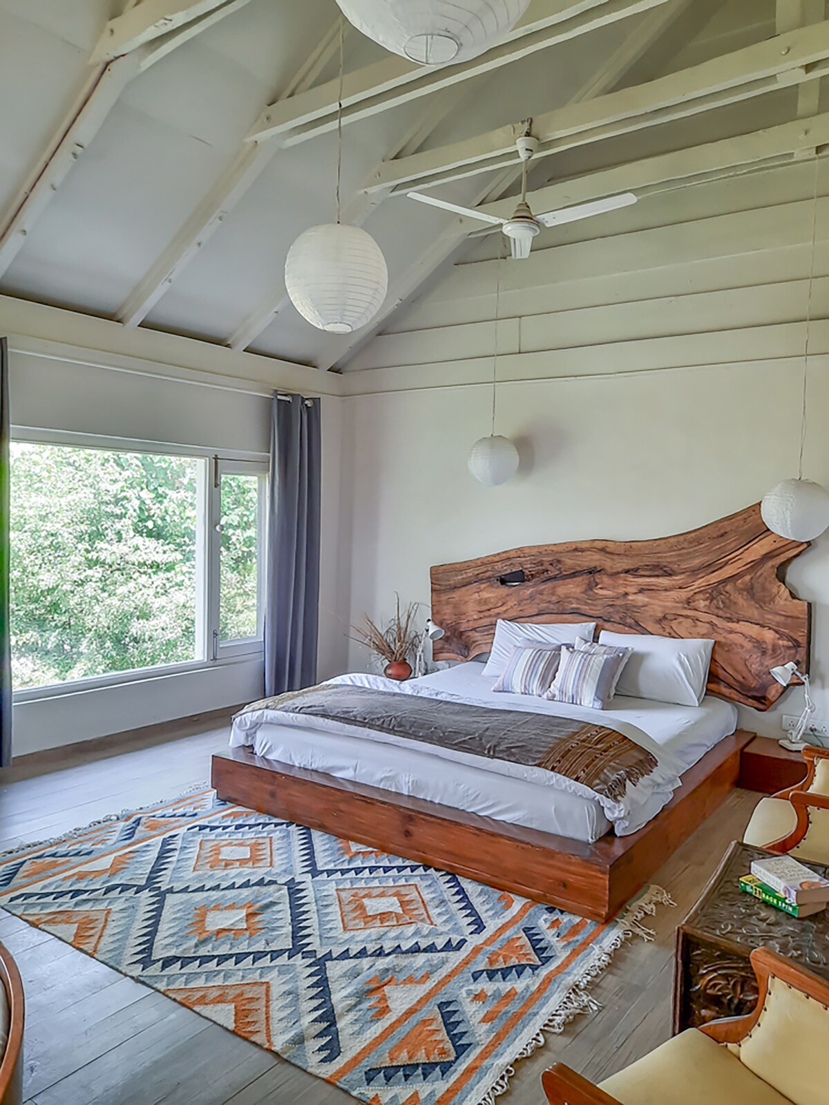 A spacious bedroom features a prominent wooden headboard above a neatly made bed with patterned pillows and a cozy throw. Natural light fills the room through large windows, while decorative pendant lights hang from a vaulted ceiling. An intricately designed rug adds warmth beneath the bed.