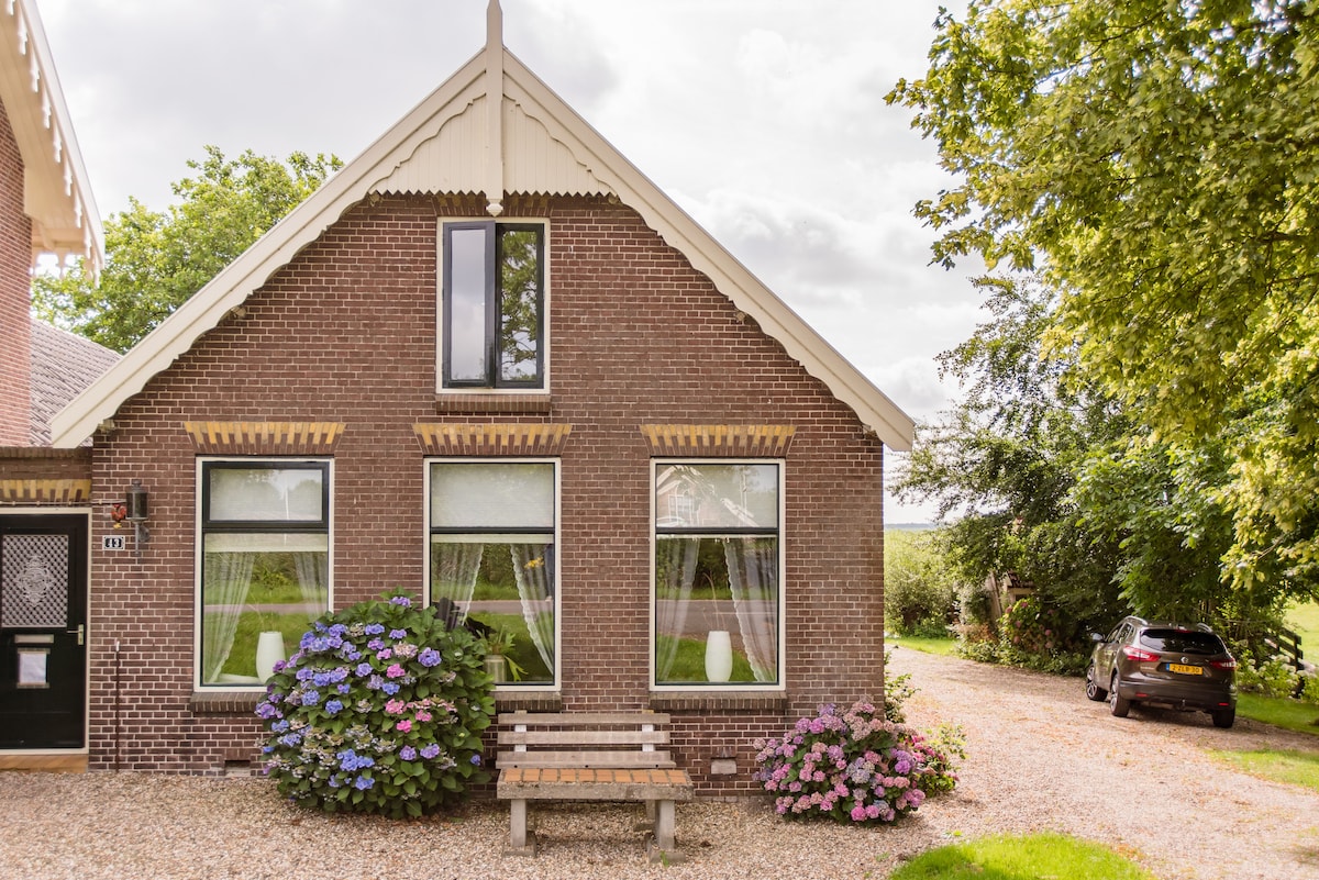 A charming house with a traditional Dutch façade is surrounded by greenery. The front features three large windows adorned with sheer white curtains, and flower beds with blooming hydrangeas provide color. A gravel driveway leads to a parked car, enhancing the welcoming atmosphere.