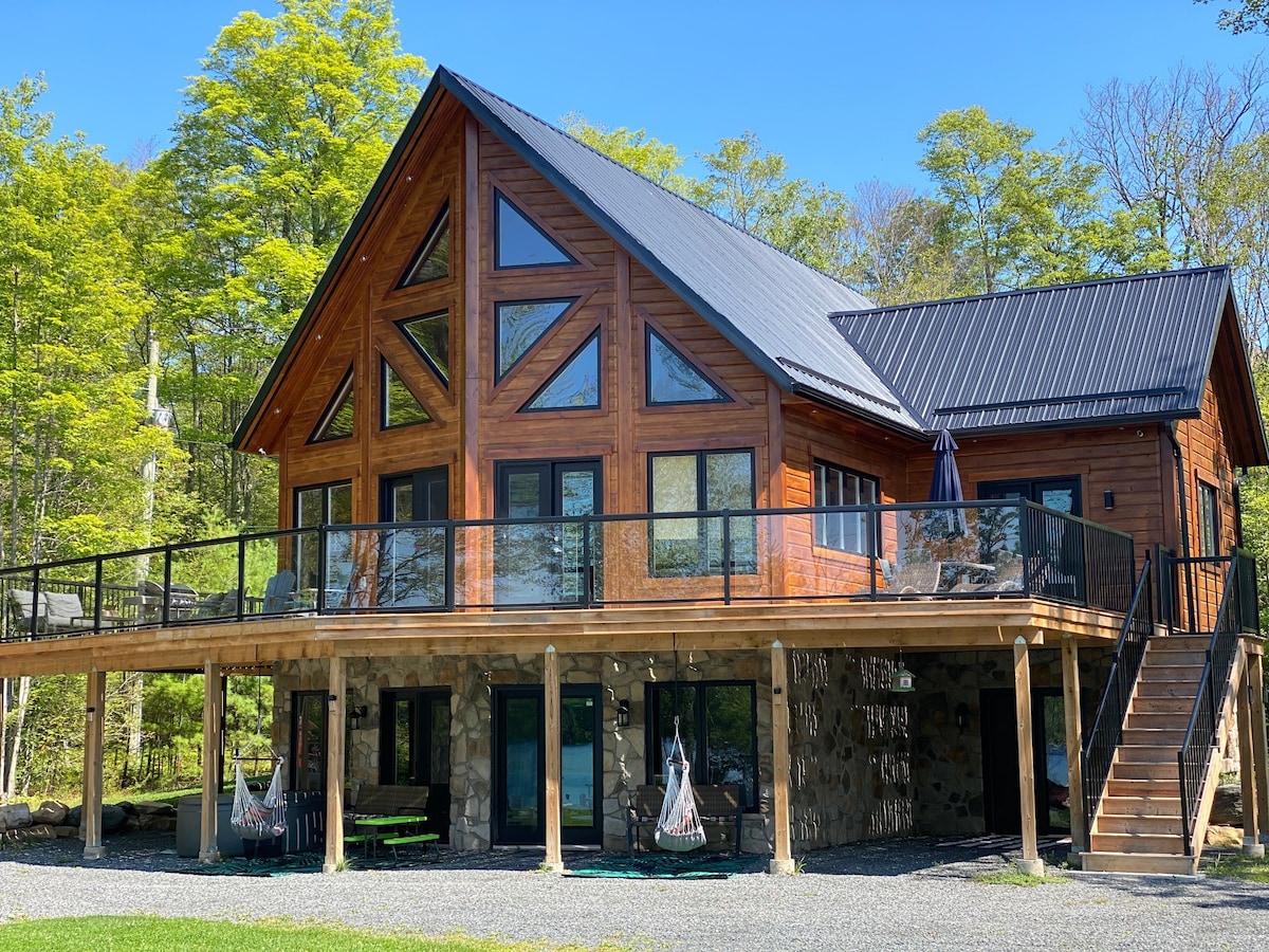 The exterior of the retreat features a rustic wooden facade with large glass windows that provide ample natural light. A spacious deck wraps around the lower level, supported by wooden beams. Vibrant greenery surrounds the property, creating a tranquil outdoor environment.