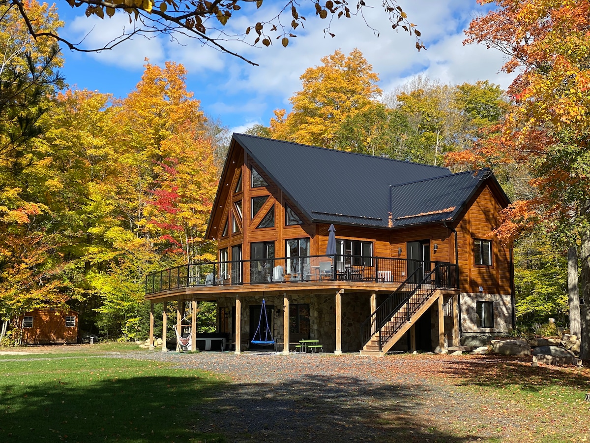 The exterior of a spacious cabin is showcased, featuring a combination of wood and stone materials. A generous deck is visible, surrounded by vibrant autumn foliage. The structure exhibits a peaked roof and large windows, allowing for plenty of natural light in an inviting setting.