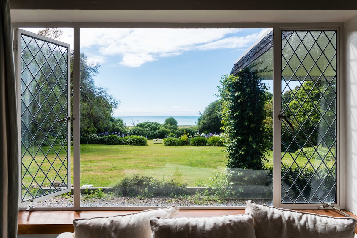 A large window with diamond-patterned glass frames a serene view of a garden leading to the coastal landscape. The lush green lawn is dotted with colorful flowers, while the calm ocean lies in the distance under a clear blue sky.