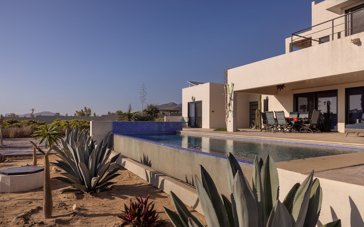 An outdoor infinity pool reflects the clear sky, surrounded by desert landscaping featuring various succulents. The spacious terrace of the house offers seating options for relaxation. The modern architecture showcases large windows and smooth surfaces, promoting seamless integration with the natural environment.
