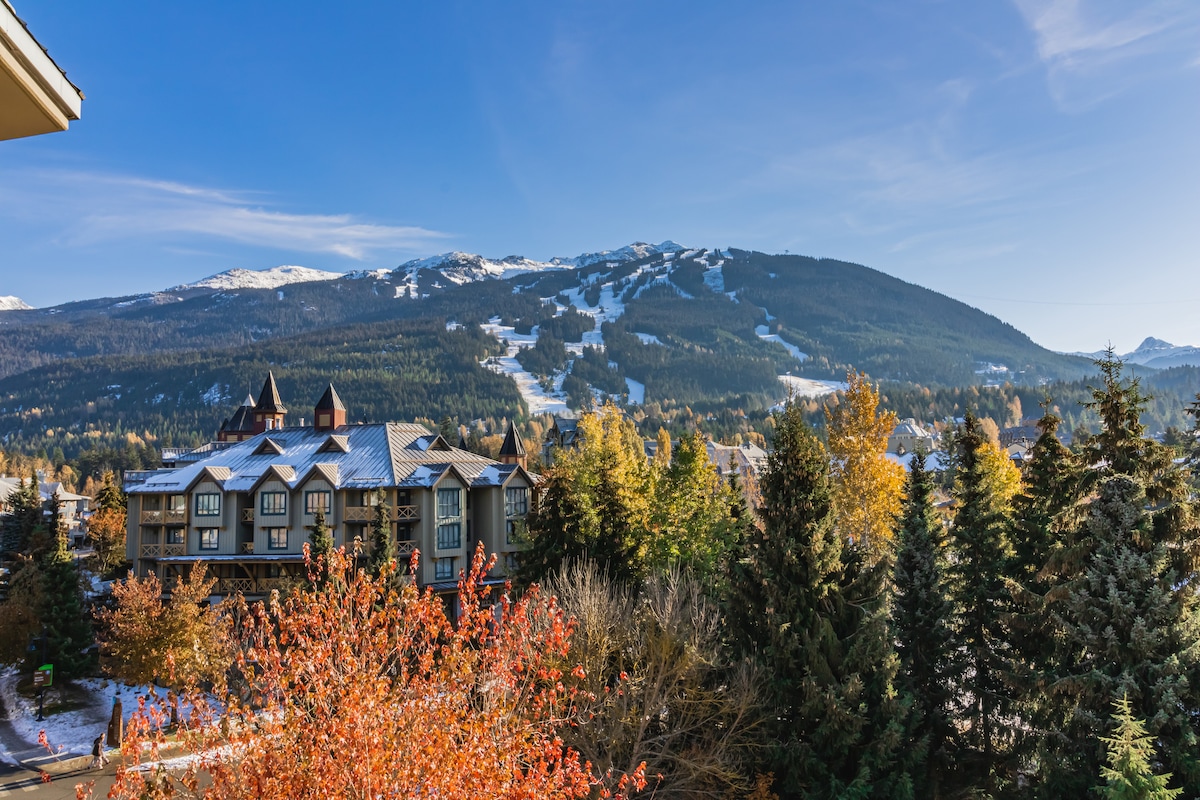 A panoramic view showcases a mountainous landscape, featuring snow-capped peaks and ski runs. Lush autumn foliage adds vibrant color, contrasting with the serene blue sky. In the foreground, a mix of evergreen trees and deciduous trees enhances the natural beauty of the scene.