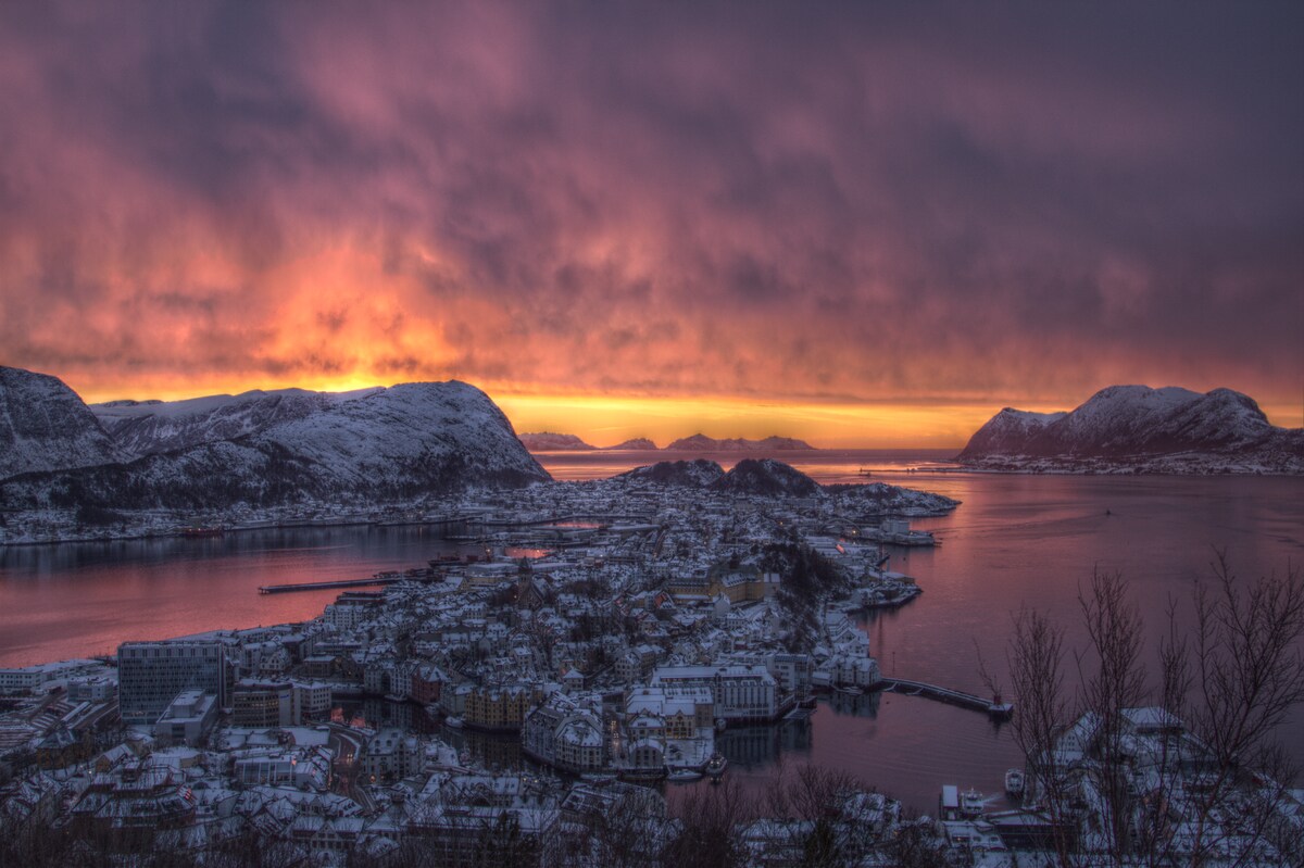 A panoramic view of a snow-covered landscape at sunset, featuring a town nestled between mountains and a fjord. The sky is filled with hues of orange and purple, reflecting on the water. Snow-capped hills frame the scene, creating a serene atmosphere.