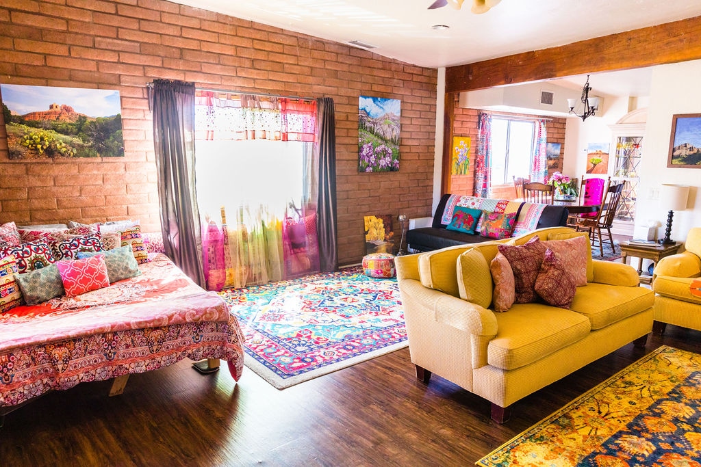 The living area features rich, colorful rugs and vibrant wall art, enhancing its bohemian style. A spacious seating arrangement includes a yellow sofa and floor seating adorned with cushions. Natural light illuminates the space, with large windows framed by flowing curtains.