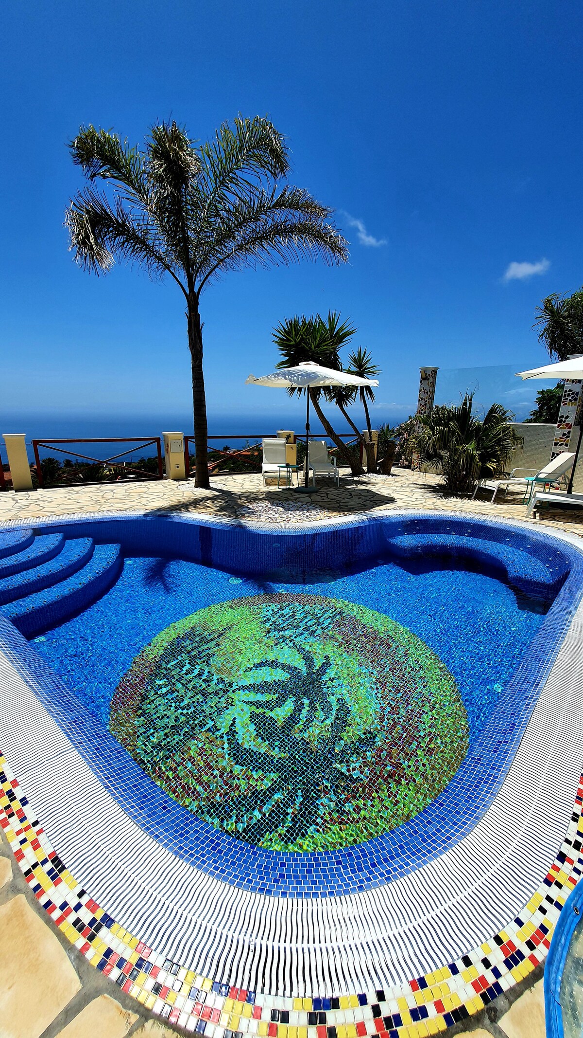 A heated pool features a vibrant mosaic design at its bottom, surrounded by a spacious patio. Sun loungers and a large umbrella are positioned nearby, with a backdrop of lush greenery and a clear blue sky. The stunning ocean view enhances the tranquil setting.