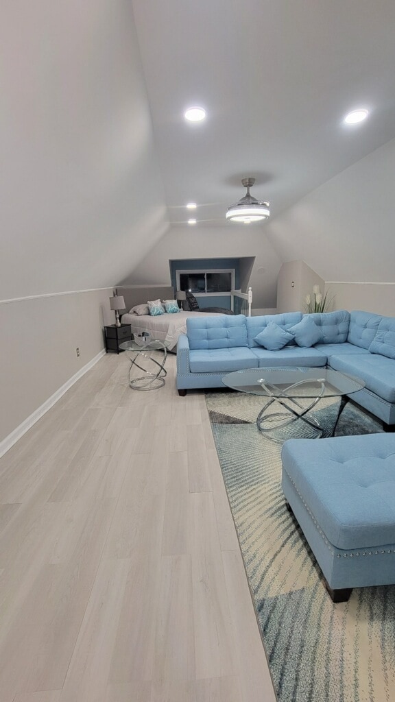 A modern living area features a light blue sectional sofa, arranged comfortably with glass side tables. Soft lighting enhances the spacious feel, while large windows allow natural light to enter. The bedroom area is visible in the background, separated by a slight partition.