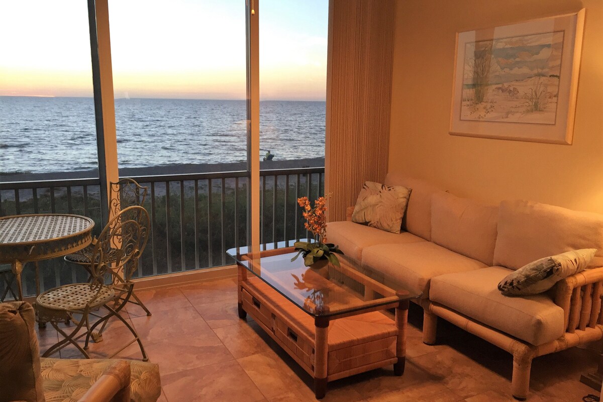 A comfortable living space features a light-colored sofa arranged around a glass coffee table. A small round dining table with iron chairs is positioned beside large windows offering unobstructed views of the water and sunset beyond.