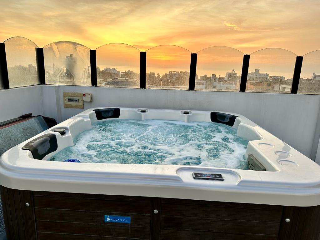 A well-appointed jacuzzi is situated on a private terrace, surrounded by protective glass panels. The bubbling water reflects the warm hues of a sunset, creating a serene ambiance. The city skyline is visible beyond, offering a panoramic view of the surrounding area.