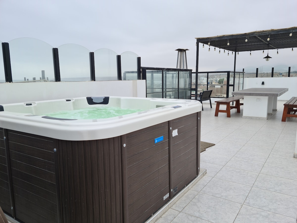 A spacious terrace area features a large jacuzzi surrounded by stylish tiles. Outdoor seating arrangements, including a large table and benches, are visible, along with a partial view of the city skyline seen through glass panels. Soft lighting hangs overhead, enhancing the area.