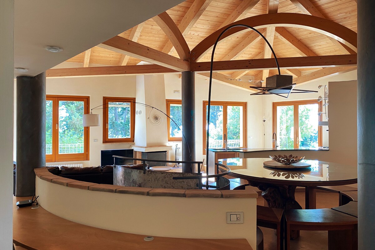 An open space kitchen and dining area is featured with wood-beamed ceilings and large windows that provide natural light. A stylish curved countertop and modern table set create a welcoming environment, surrounded by views of the garden.