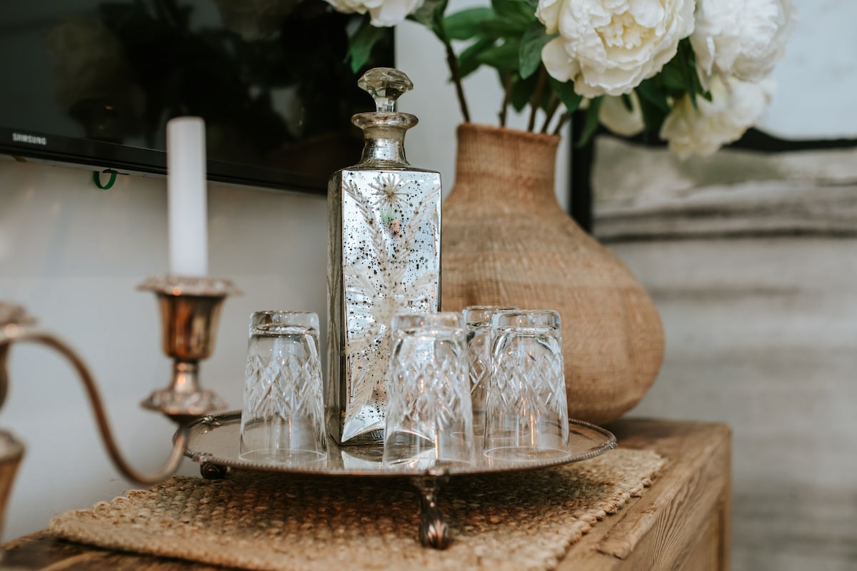An elegant display features a sparkling silver decanter alongside six intricately designed glass tumblers. A candle stands beside them on a woven tray, framed by a large woven vase adorned with white peonies, contributing to a refined setting.