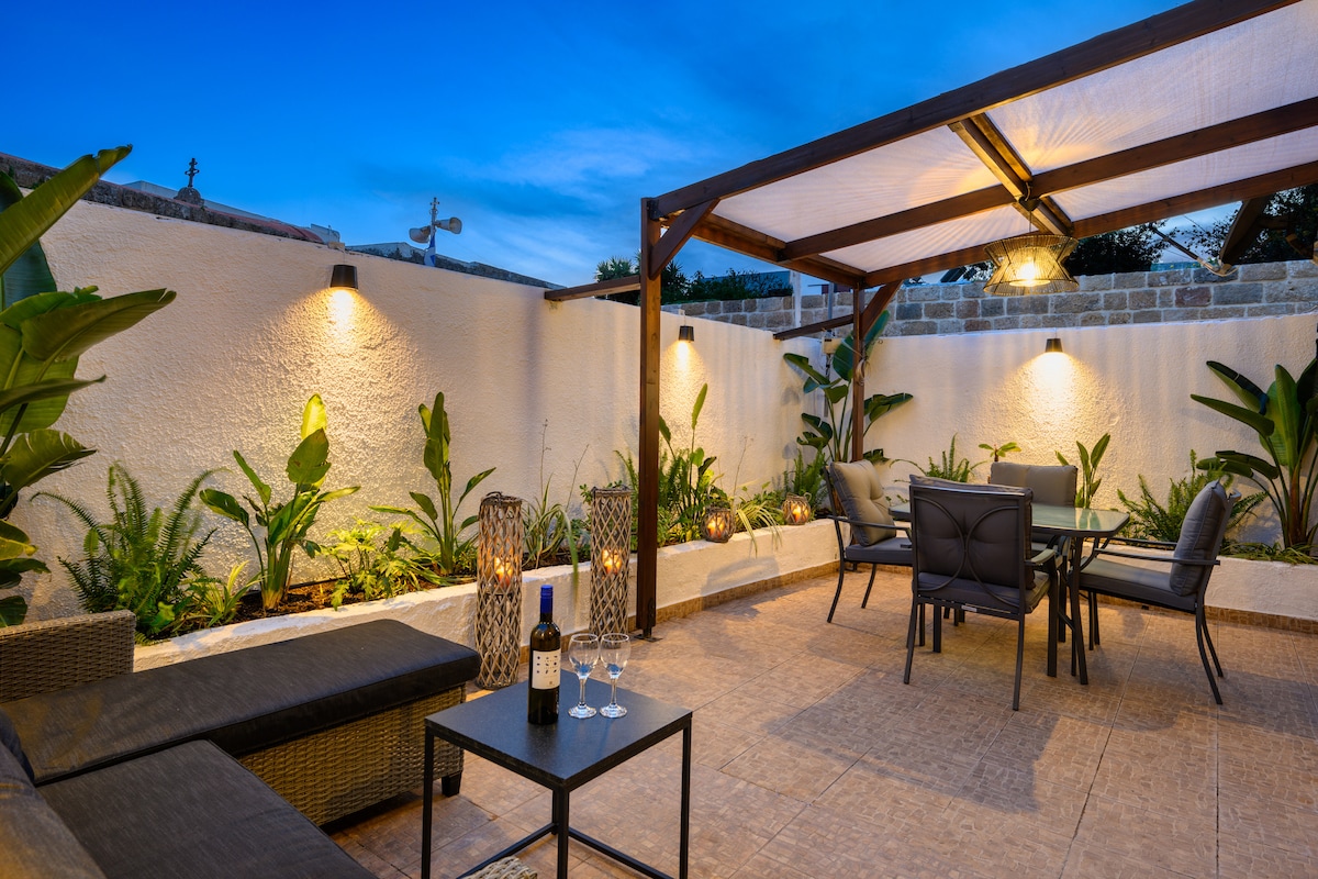 An intimate outdoor space is highlighted by a covered seating area, featuring a dining table surrounded by chairs. Lush greenery and ambient lighting create a serene environment, with a sleek table set for wine, enhancing the overall tranquility of the area.