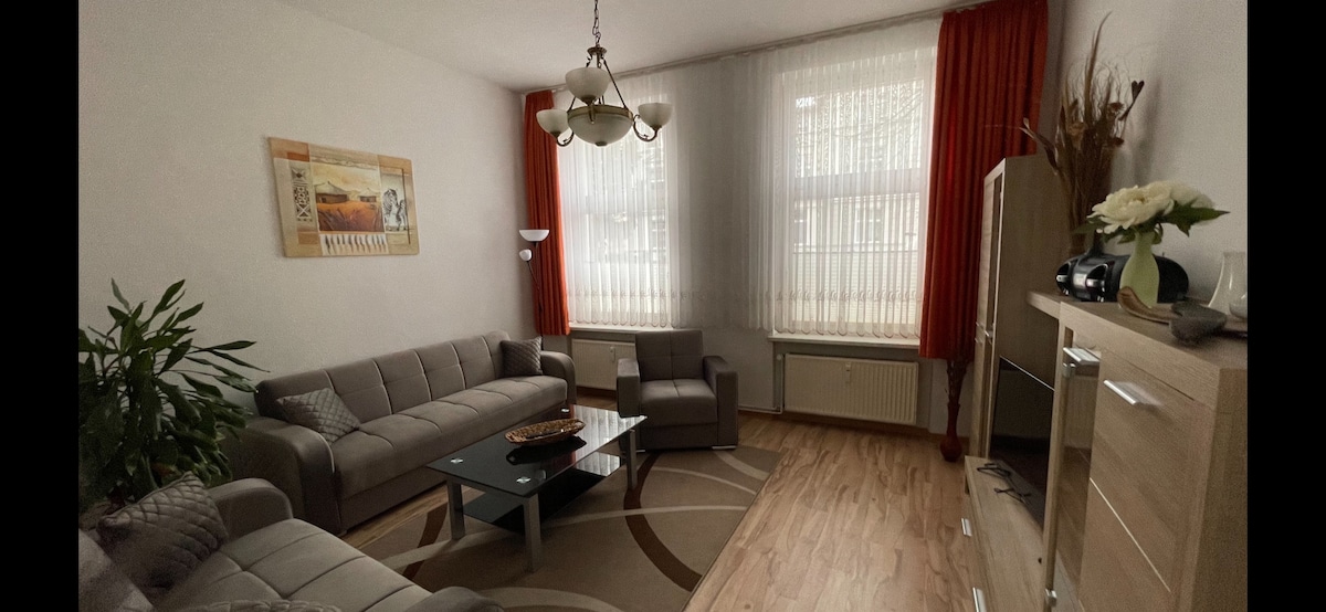 A spacious living room features a light-colored sofa set arranged around a coffee table. Large windows are dressed with sheer curtains, allowing natural light to fill the space. Decorative elements and light-colored walls create a calm setting, complemented by a wooden sideboard.