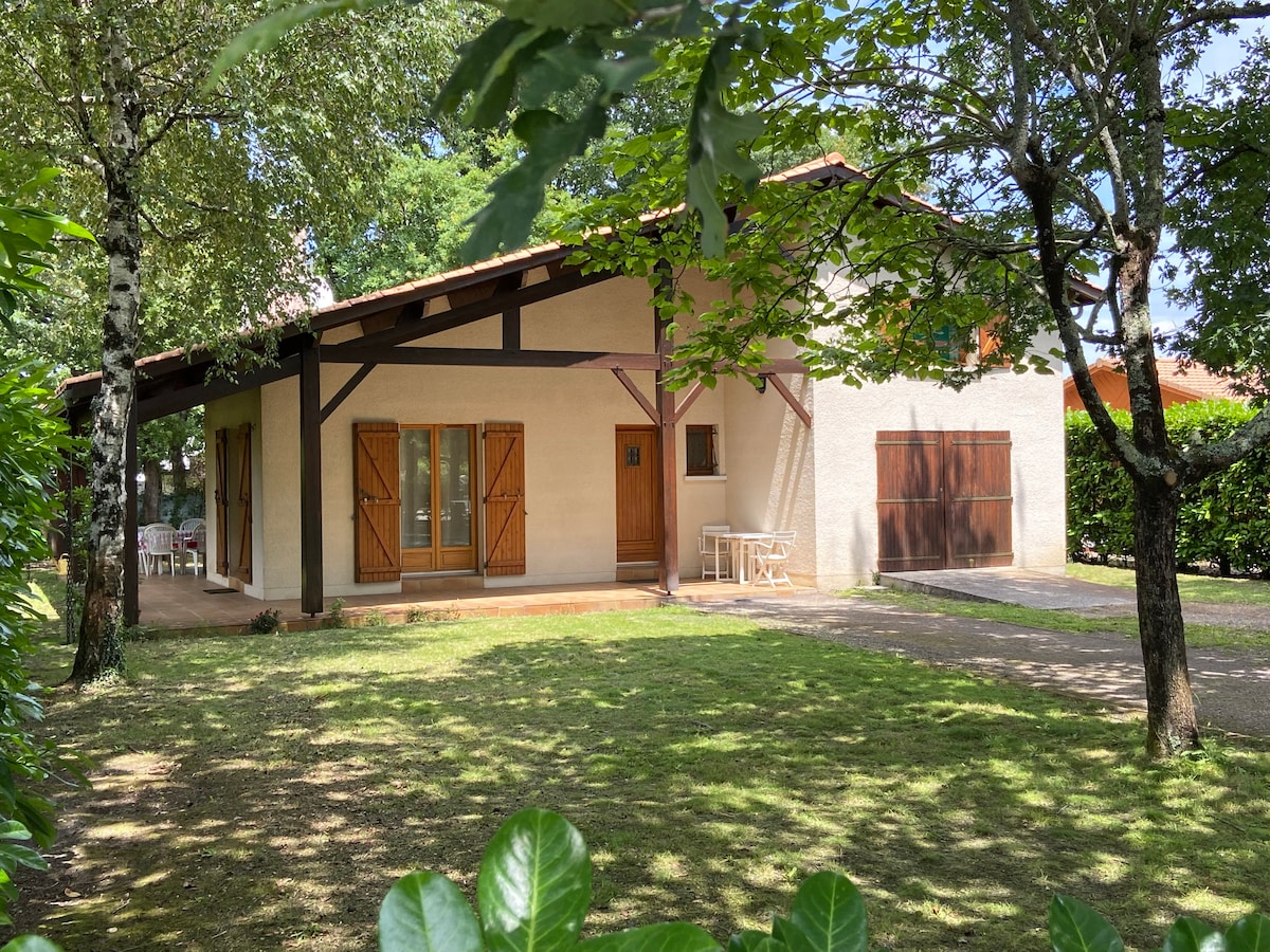 A single-story house is nestled among trees in a landscaped yard. The exterior features light-colored walls and brown shutters, with a porch that includes a small table and chairs. Grass covers the ground, and a garage door is visible to the right.