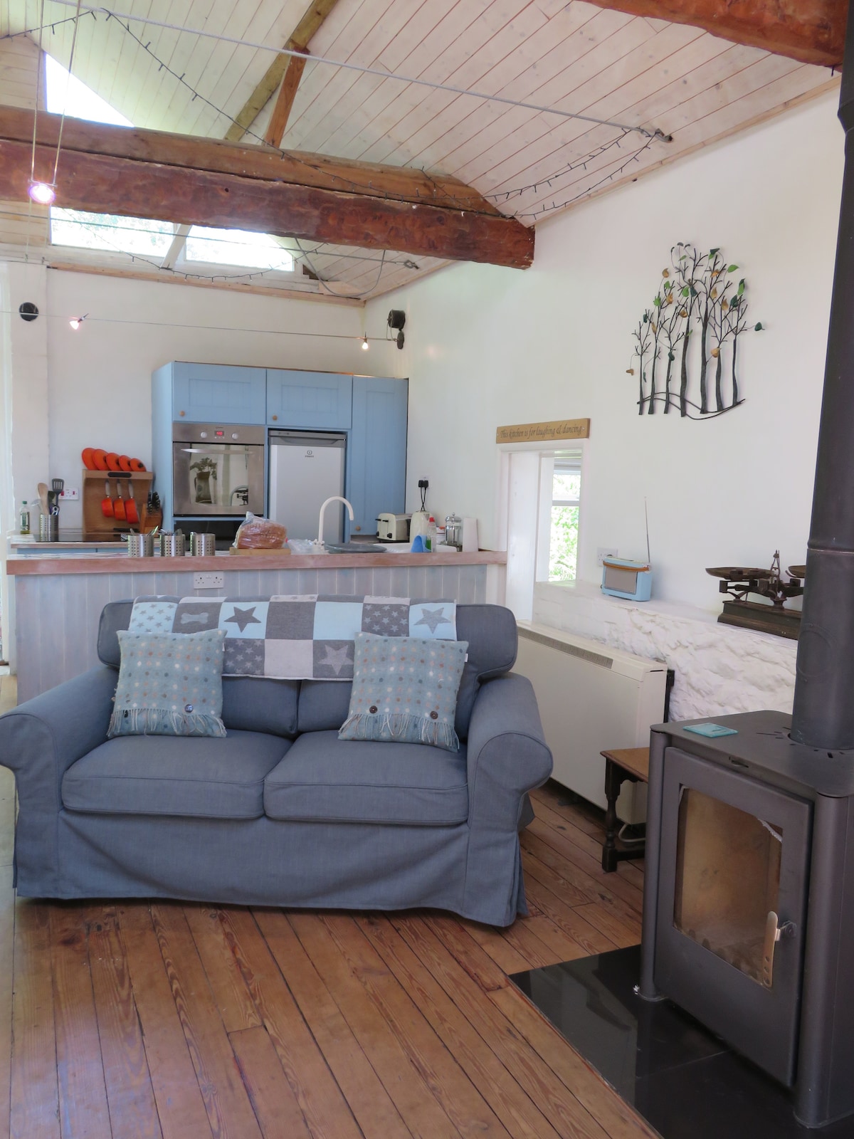 A spacious living area is featured, with a comfy blue sofa adorned with decorative pillows. A log burner is situated beside the sofa, while a modern kitchen area with light cabinetry is visible in the background, complemented by wooden flooring and natural light from the windows.