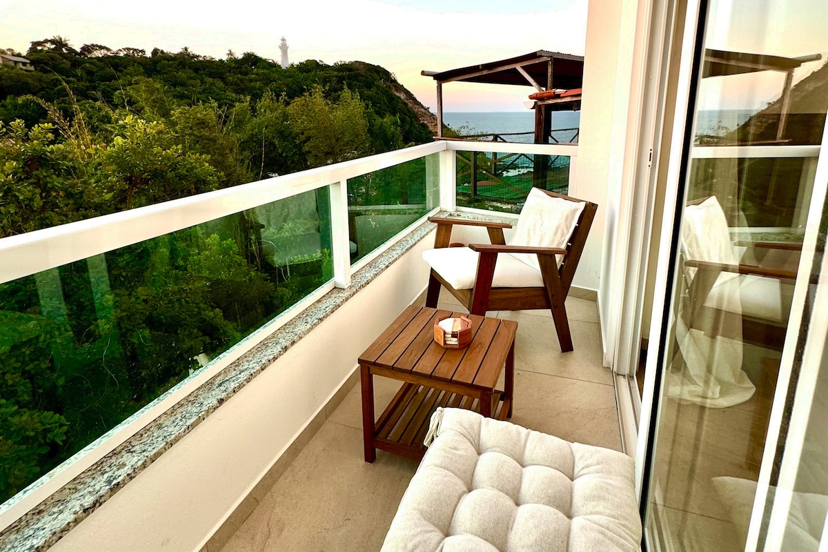 An inviting balcony features comfortable seating and a small wooden table, perfect for relaxation. Glass railings provide unobstructed views of the lush surroundings and ocean. The gentle glow of a candle adds a soft ambiance as the sun sets on the horizon.