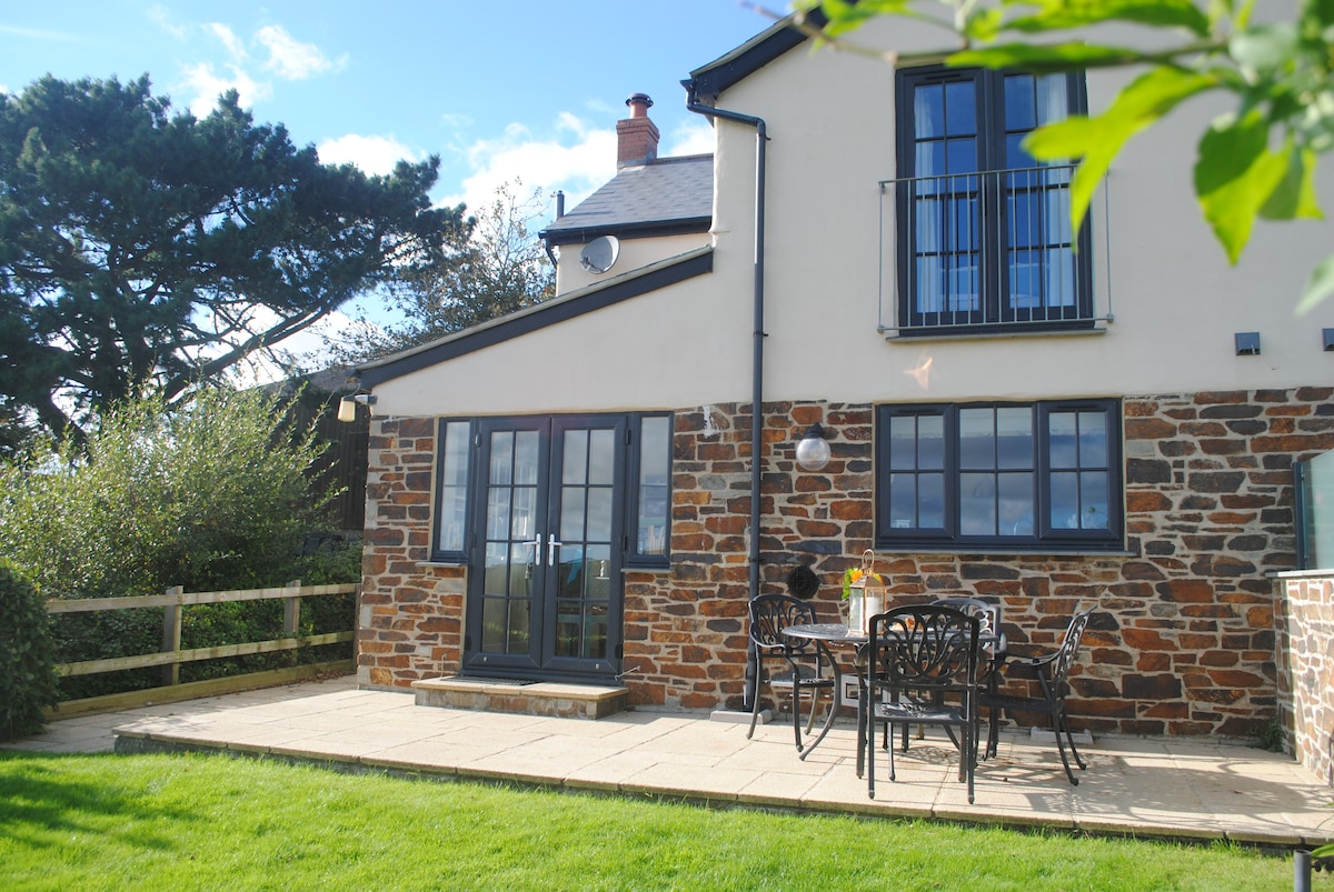 The exterior of the semi-detached cottage features a private patio area with outdoor seating for four. Stone walls and large windows reflect the natural surroundings. A landscaped garden with grass and wooden fencing provides a tranquil atmosphere, complemented by nearby trees.