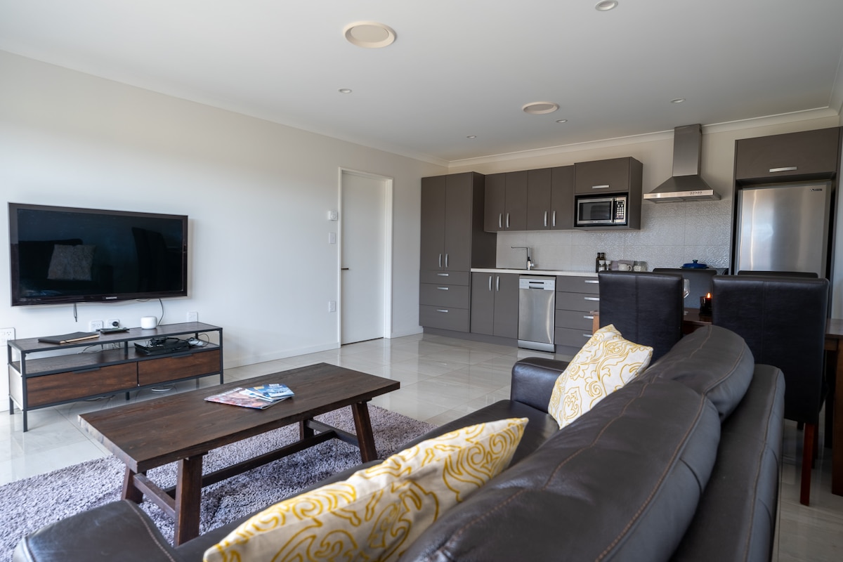 A modern open-plan living area features a comfortable dark sofa with decorative throw pillows. A polished wooden coffee table sits in front, and a flat-screen TV is mounted on the wall. The kitchen area is equipped with stainless steel appliances and sleek cabinetry.