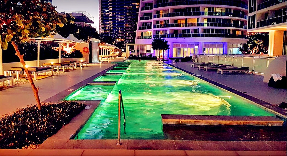 An outdoor swimming area is illuminated with soft green lighting, showcasing multiple shallow pools arranged in a row. Surrounding the pools are lounge chairs and shaded seating areas, framed by trees and modern architectural features of the building.