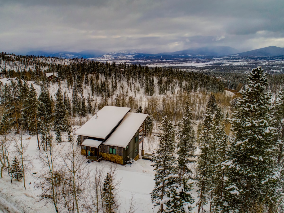 An aerial view showcases a modern mountain home surrounded by a snowy landscape. Towering evergreens frame the property, with distant mountain ranges visible under a cloudy sky. The snow-covered roof and exterior reflect the serene beauty of winter in Grand County.