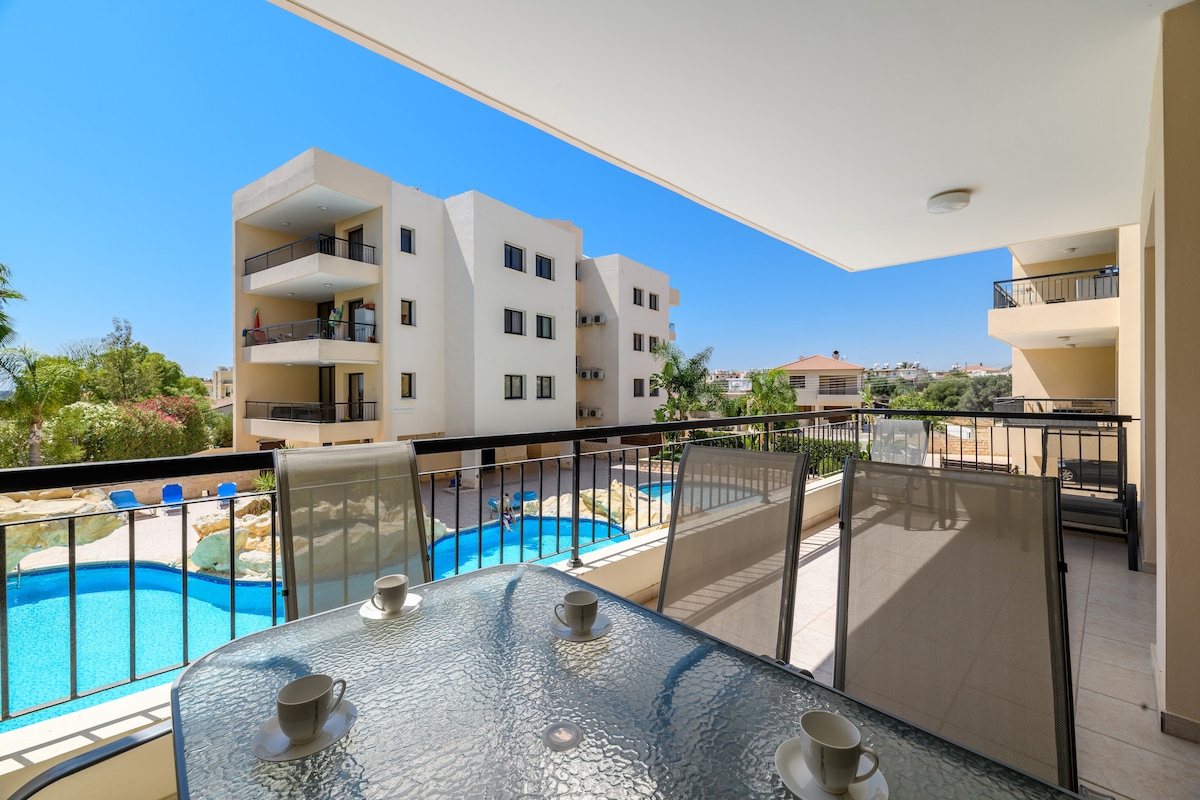An outdoor veranda features a glass table with four place settings, overlooking two inviting swimming pools surrounded by landscaped greenery. A bright blue sky complements the serene scene, with neighboring apartment buildings partially visible in the background.
