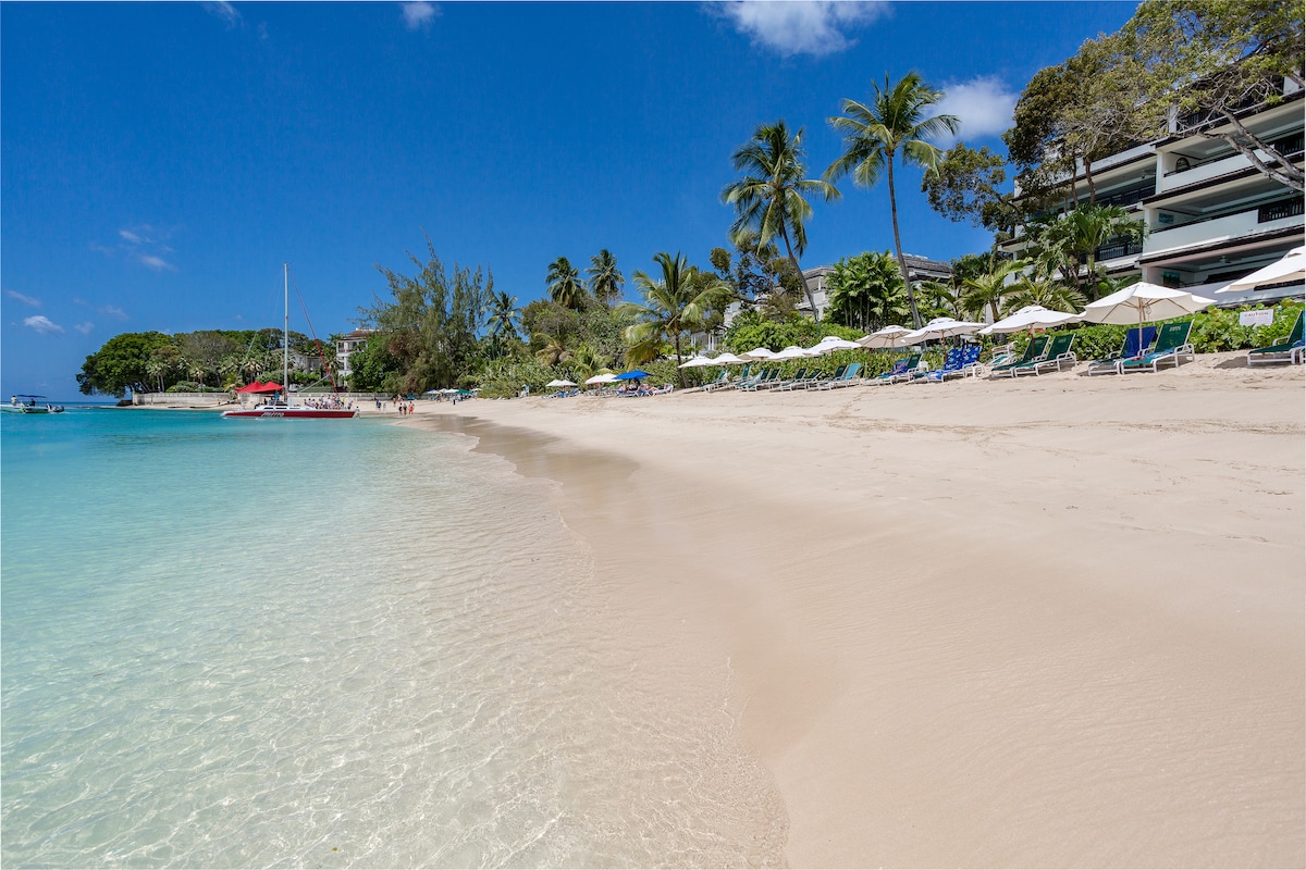 The image captures a serene beach scene, showcasing soft sand meeting clear turquoise waters. Sun loungers are arranged under umbrellas along the shore, with palm trees providing gentle shade. A few boats are visible in the distance, contributing to the tranquil coastal atmosphere.