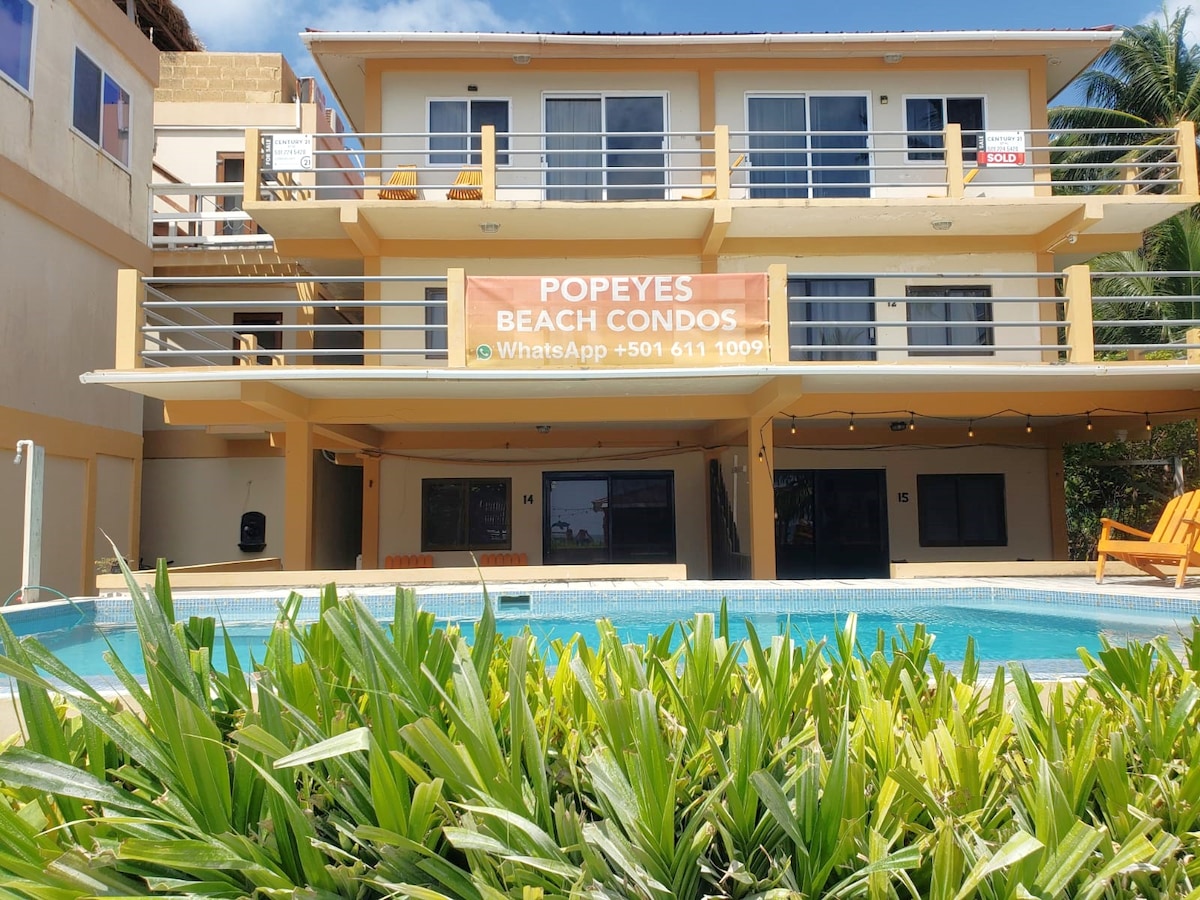 A three-story building with a bright banner reading 'Popeyes Beach Condos' is observed. Balconies with chairs overlook a shared pool, surrounded by lush greenery. The structure's exterior is painted in neutral tones, contributing to a relaxed seaside ambiance.